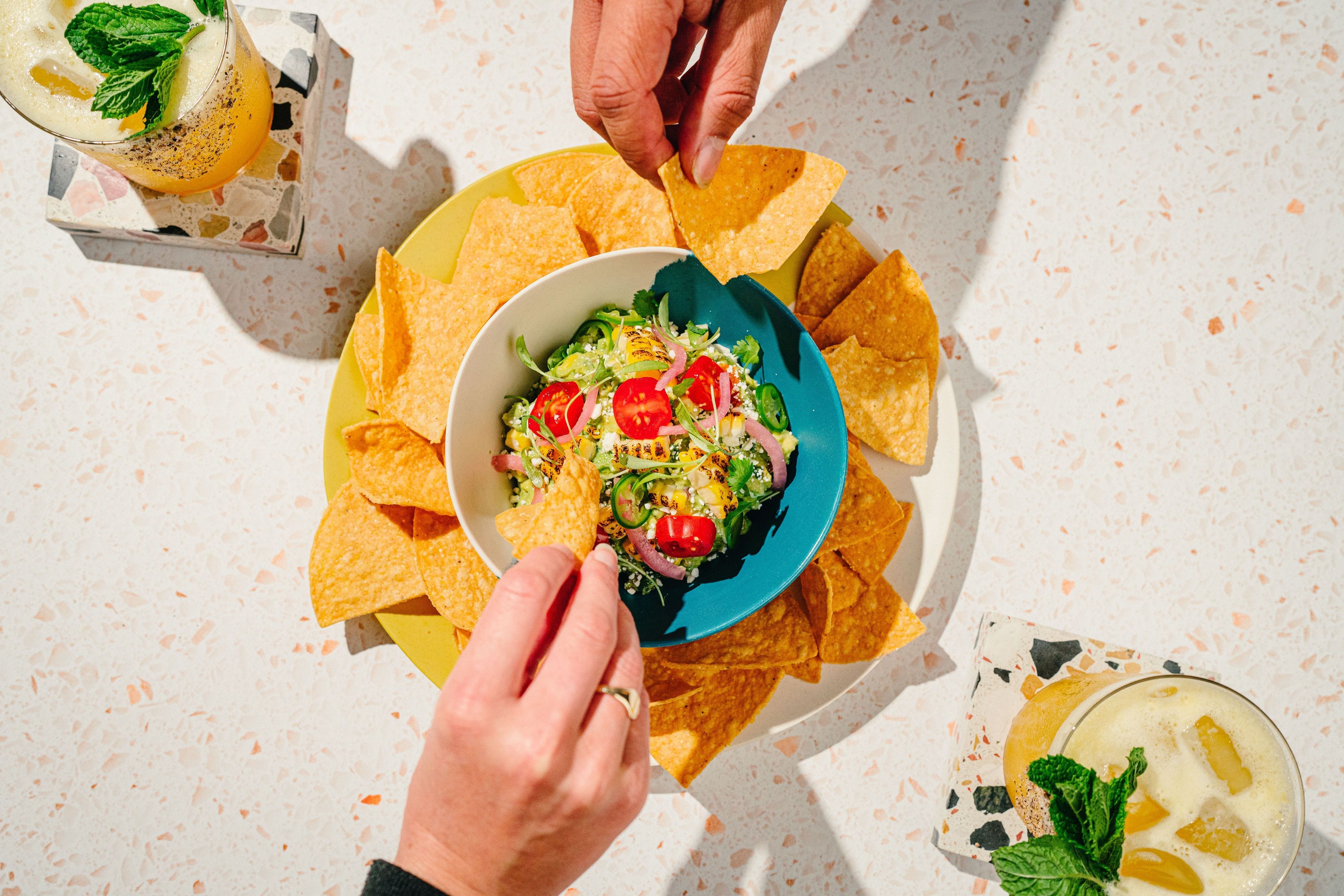 Two hands reaching for tortilla chips around a bowl of grilled corn salad and cocktails.